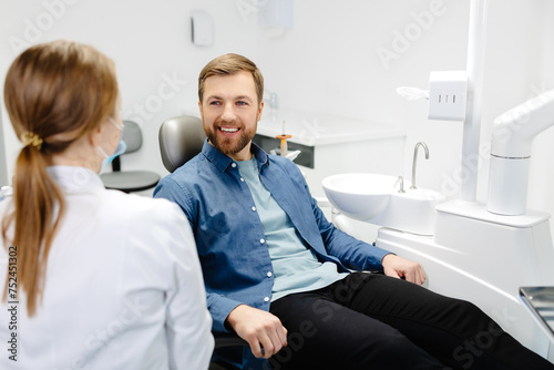 Blonde female dentist in dental office talking with male patient and preparing for treatment. Handsome bearded man in dentist chair looking at his doctor with smile