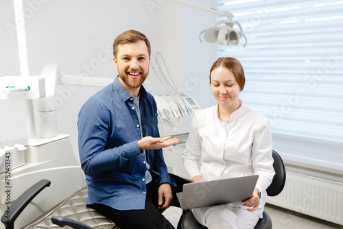 A handsome young man is talking to a female doctor at a dental appointment in a bright  beautiful office. The dentist explains to the patient and shows everything on the laptop