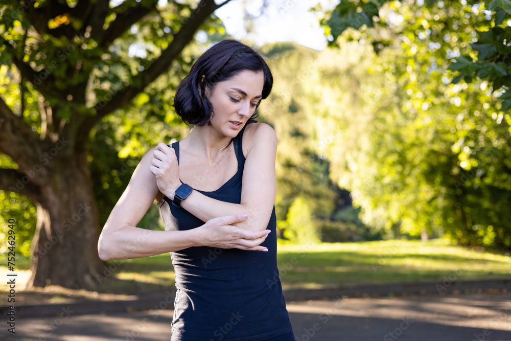 Young sporty woman standing outside in park and holding hand on elbow, injured, feeling severe pain Young sporty woman standing outside in park and holding hand on elbow, injured, feeling severe pain