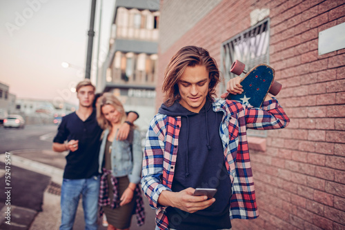 Young man with skateboard checking phone with friends in background photo