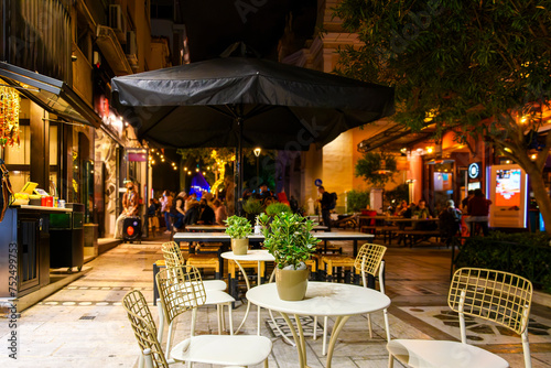 Fototapeta Naklejka Na Ścianę i Meble -  A fresh plant sits on a table for two at a sidewalk cafe in the Monastiraki district of downtown Athens, Greece at night, with colorful cafes and shops blurred behind.