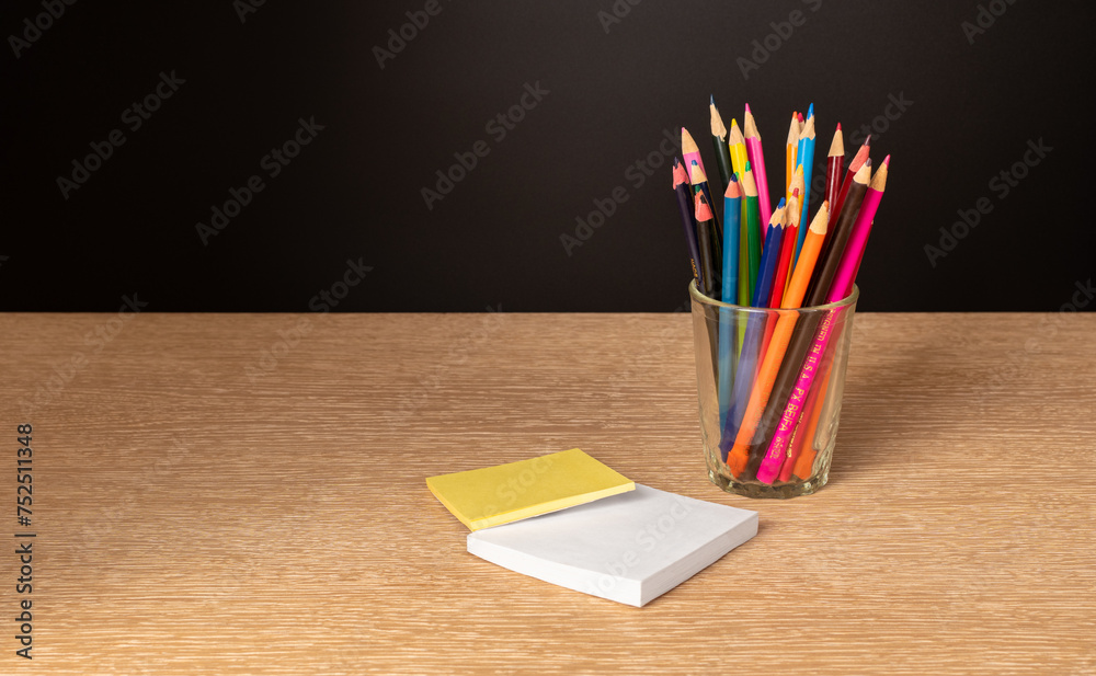 A glass with colored pencils and stickers on a wooden table and a black background