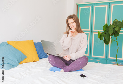 Happy young woman sitting on the bed and working with computer at home