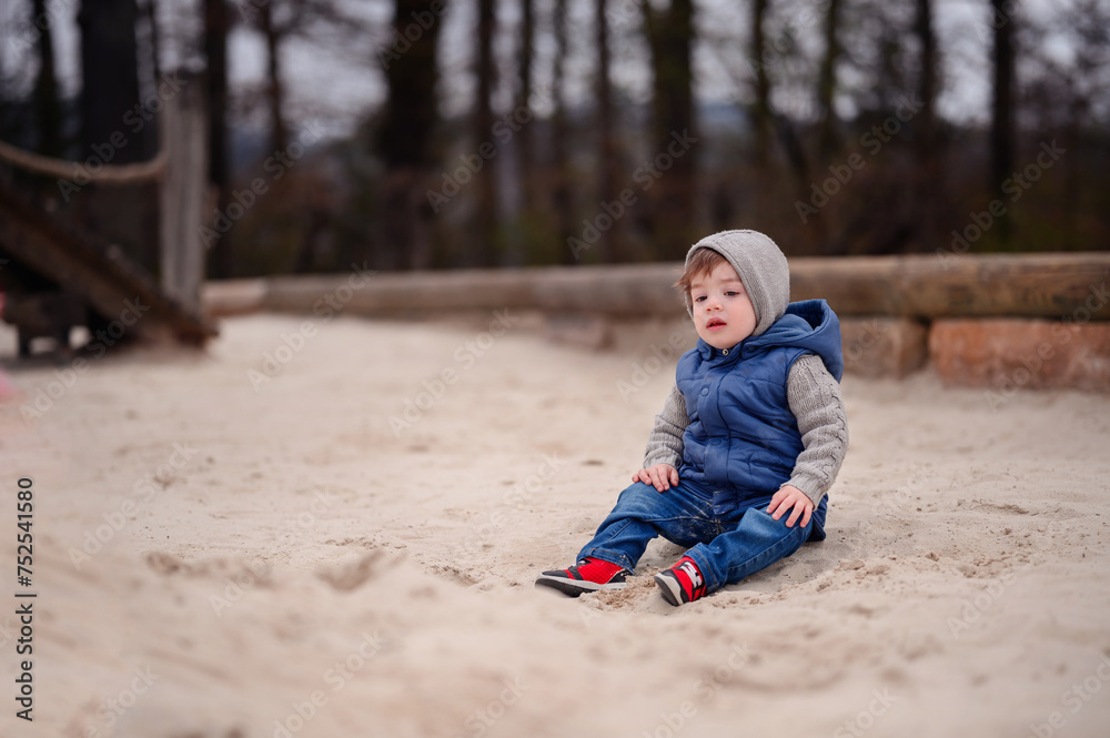 A young child dressed warmly in a vest and hat sits thoughtfully in the sand of a playground, capturing a moment of childhood tranquility and exploration A young child dressed warmly in a vest and hat sits thoughtfully in the sand of a playground, capturing a moment of childhood tranquility and exploration