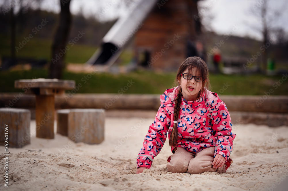A cheerful girl with glasses plays in the sand, her bright pink jacket adding a pop of color to the playful scene at the park A cheerful girl with glasses plays in the sand, her bright pink jacket adding a pop of color to the playful scene at the park