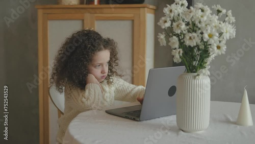 Sweet cute curly hair Bored School girl child using laptop online educational lesson course at home. Distance learning course remote video conference. Schoolgirl study does school homework in kitchen photo