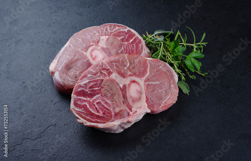 Raw beef leg slice with cross-section marbling with herbs and spices as ossobuco offered close-up on a black design board with text space