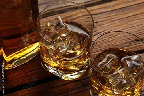 Whiskey with ice cubes in glasses and bottle on wooden table, closeup