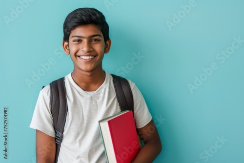 Smiling Indian boy student in casual clothes holding a backpack and books against a plain light blue background