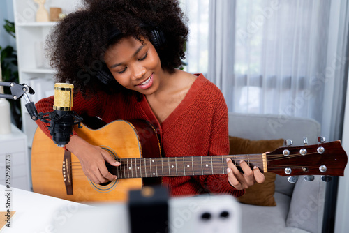 Host channel of beautiful African woman influencer singing with playing guitar in broadcast studio on smartphone. Time slot of music blogger on live social media. Concept of audio creator. Tastemaker. photo