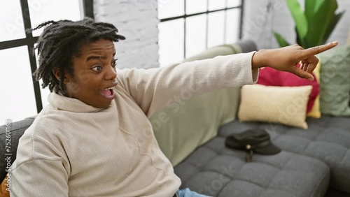 Shocked young black woman with dreadlocks, sitting cozy on sofa home, pointing ahead in surprise! open mouth, amazed expression discovering something fun forward. photo