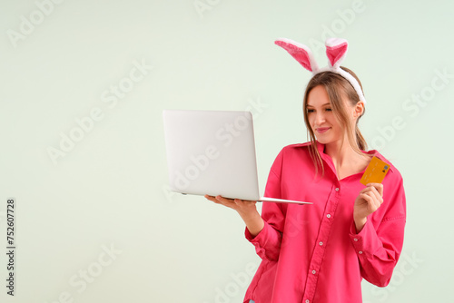 Young woman in bunny ears with laptop and credit card on light background. Easter Sale