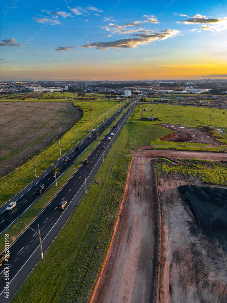 Imagem aérea da cidade de Paulínia durante por do sol com tráfego e construção de condomínio residencial. 2024.