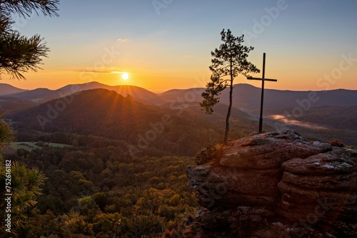 Sunrise on the Roetzenfelsen in the Palatinate Forest with bright sunshine and the lone pine tree and the summit cross on the red rock
