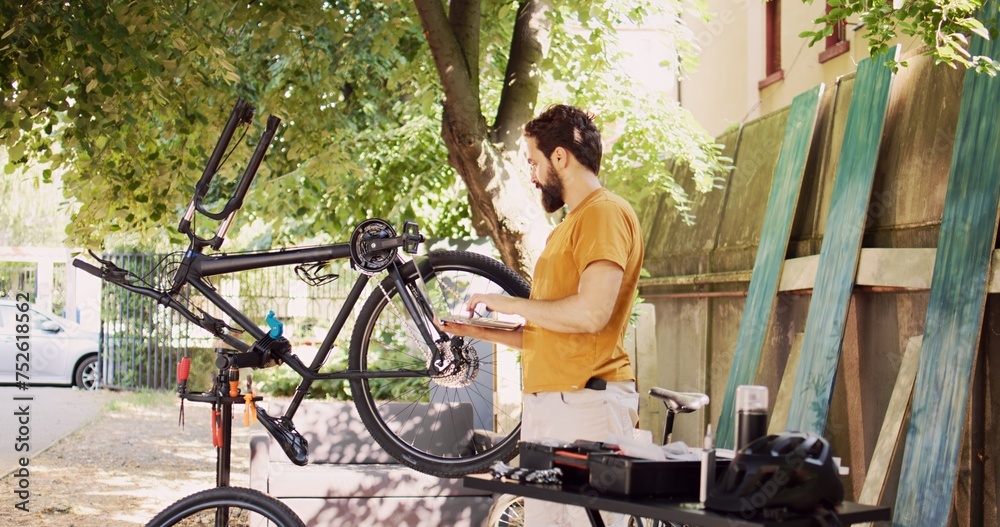 Obraz premium Male cyclist holding personal computer and inspects bike pedal and crank arm for damages in home yard. Active caucasian man enjoys browsing internet, fixing and maintaining modern bicycle.