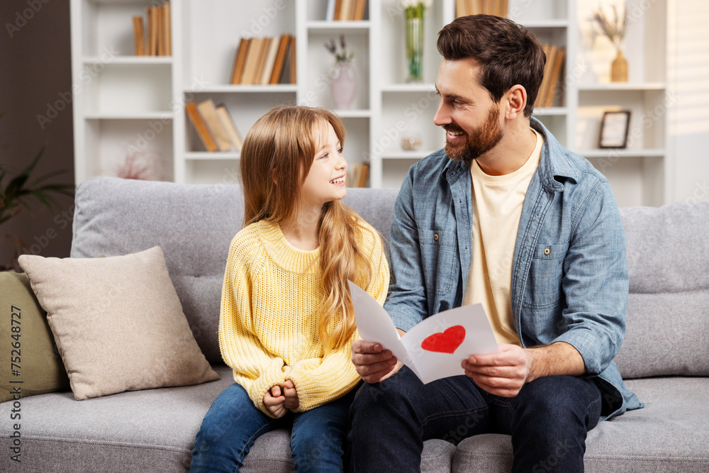 Smiling father holding a card from his little daughter, both sitting on sofa, looking at each other Smiling father holding a card from his little daughter, both sitting on sofa, looking at each other
