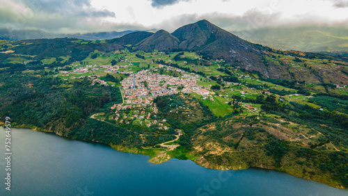 Aerial view of Guatavita town nestled by the tranquil Tomine Reservoir, surrounded by lush green hills under a cloudy sky