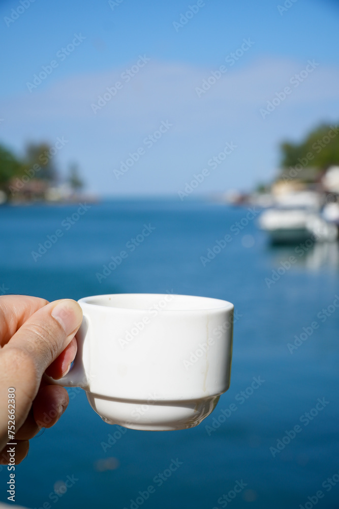 Obraz premium Man holds a white coffee cup in his hand on the background of the sea and boat.