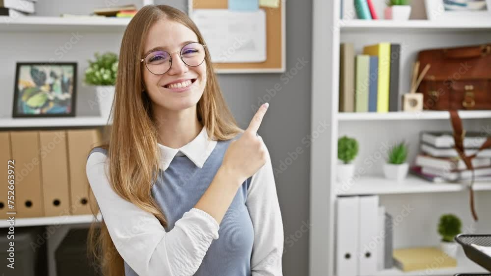 Cheerful young blonde business woman lighting up the office, confidently pointing to the side, her warm smile capturing the camera. Cheerful young blonde business woman lighting up the office, confidently pointing to the side, her warm smile capturing the camera.