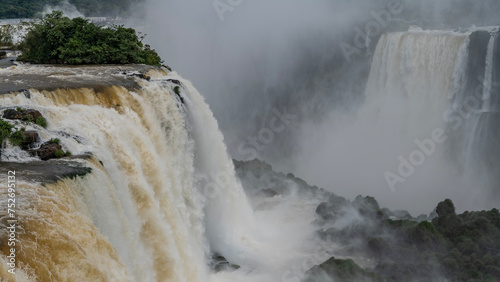 The powerful foaming streams of the waterfall collapse in the gorge. Poor visibility due to splashes, fog. Green vegetation on the rocks. Iguazu Falls. Brazil