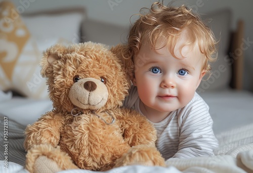 Smiling Baby With Blue Eyes Next to a Brown Teddy Bear on a Soft Bed © Olena Rudo