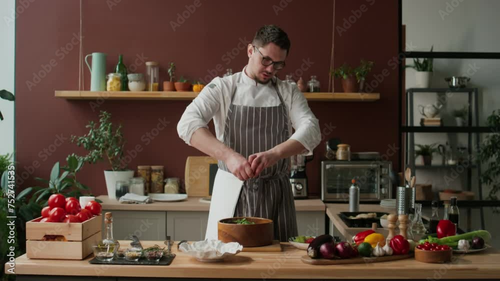 Medium shot of Caucasian young male chef in workwear and eyeglasses squeezing grapefruit juice into salad while recording cooking show in domestic kitchen Medium shot of Caucasian young male chef in workwear and eyeglasses squeezing grapefruit juice into salad while recording cooking show in domestic kitchen