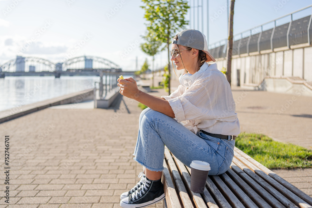 Fototapeta premium Young Hispanic hipster girl browsing mobile phone while sitting in street with takeaway beverage