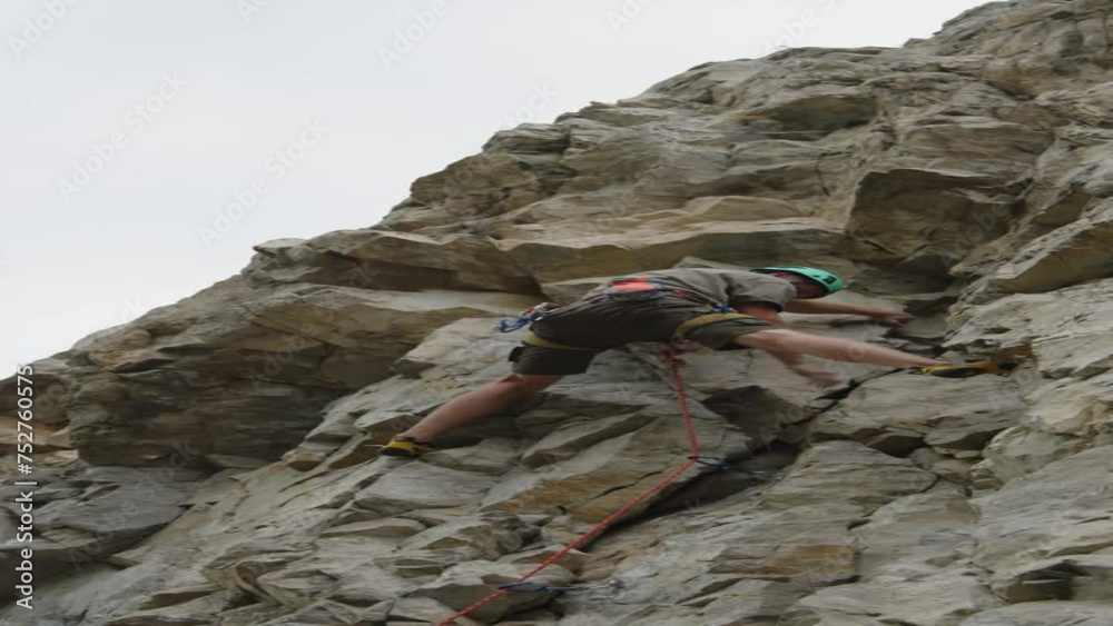 Vertical low angle shot of mountain climber wearing helmet climbing rocky cliff with rope on summer day Vertical low angle shot of mountain climber wearing helmet climbing rocky cliff with rope on summer day