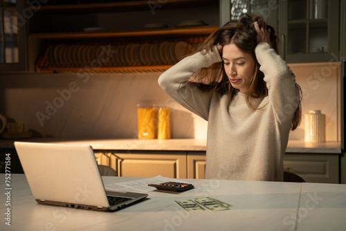 A young woman sets at her kitchen table in the evening, her brows furrowed in concentration as she works on a budget. Surrounded by papers and a laptop, she uses a calculator to manage your finances