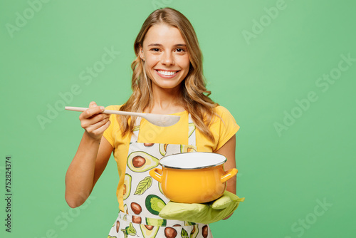 Young fun housewife housekeeper chef cook baker woman wear apron yellow t-shirt hold in hand pot pan try soup with wooden spoon isolated on plain pastel green background studio. Cooking food concept.