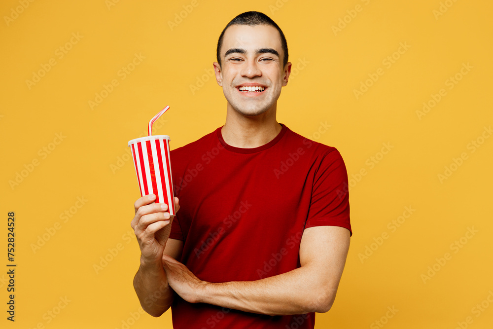 Young smiling happy middle eastern man wear red t-shirt casual clothes hold cup of soda pop cola fizzy water look camera isolated on plain yellow orange background studio portrait. Lifestyle concept. Young smiling happy middle eastern man wear red t-shirt casual clothes hold cup of soda pop cola fizzy water look camera isolated on plain yellow orange background studio portrait. Lifestyle concept.