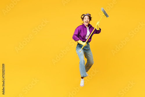 Full body excited young fun woman wear purple shirt casual clothes do housework tidy up jump high hold brush broom jump high isolated on plain yellow background studio portrait. Housekeeping concept.