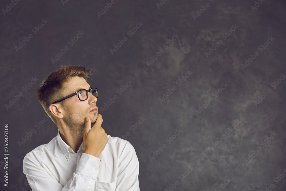 Serious millennial Caucasian man in glasses on black studio background look at blank empty copy space thinking making decision. Pensive young male consider sale deal or promotion offer. Copyspace. Serious millennial Caucasian man in glasses on black studio background look at blank empty copy space thinking making decision. Pensive young male consider sale deal or promotion offer. Copyspace.