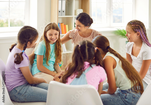 Friendly young school psychologist woman having conversation with a group of cute children girls sitting in a circle in classroom. Psychological support for kids and mental health concept.