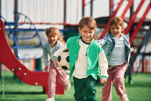 Boy is in green clothes, with soccer ball. Kids are having fun on the playground photo