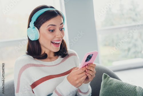 Photo of charming positive person sit chair use smart phone listen new single song headphones spend weekend house indoors