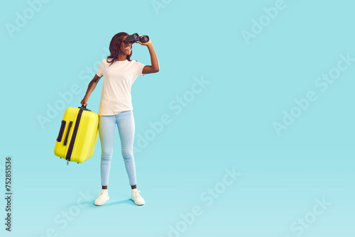 Full length portrait of happy smiling african american woman tourist in casual clothes with suitcase looking through binoculars on studio blue background. Summer holidays trip and vacation concept.