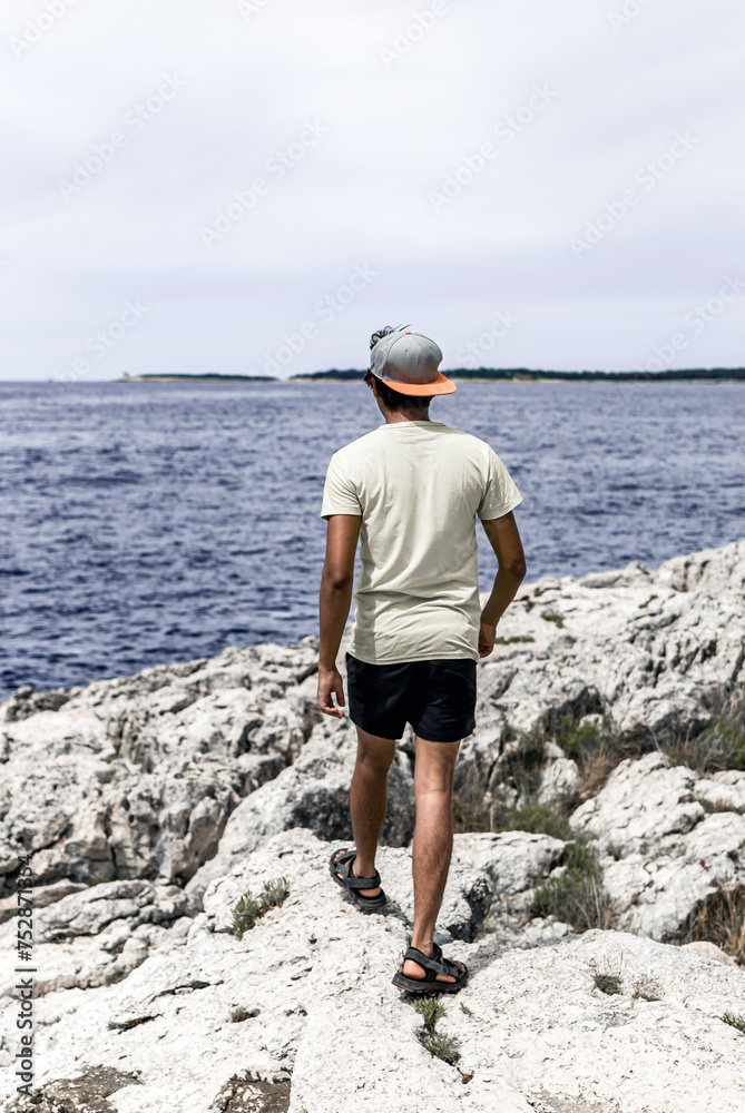 Young man with tan skin walking on the rocky beach. He is wearing shorts, t-shirt and a cap.