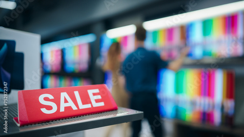 Close Up of a Red Sale Sign in a Home Electronics Department Store with a Range of Modern Smart TV Sets. Shoppers Explore Discounted Home Appliances in a Busy Retail Storefront Showroom