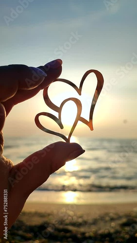 Woman holding in fingers black outline in shape of two hearts against background of sun at dawn sunset on sea. Concept love infatuation romantic Valentine's Day mood emotions relationship Vertical