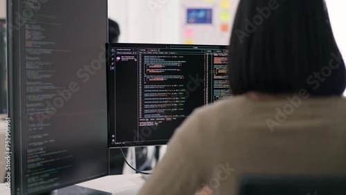A female developer is observed from behind, scrutinizing a complex codebase on her monitor within a modern technological workspace, indicative of her problem-solving capabilities.