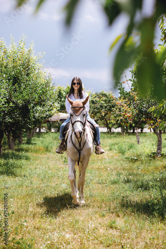 A woman with sunglasses enjoying a horseback ride amidst the blossoming orchard on a sunny day