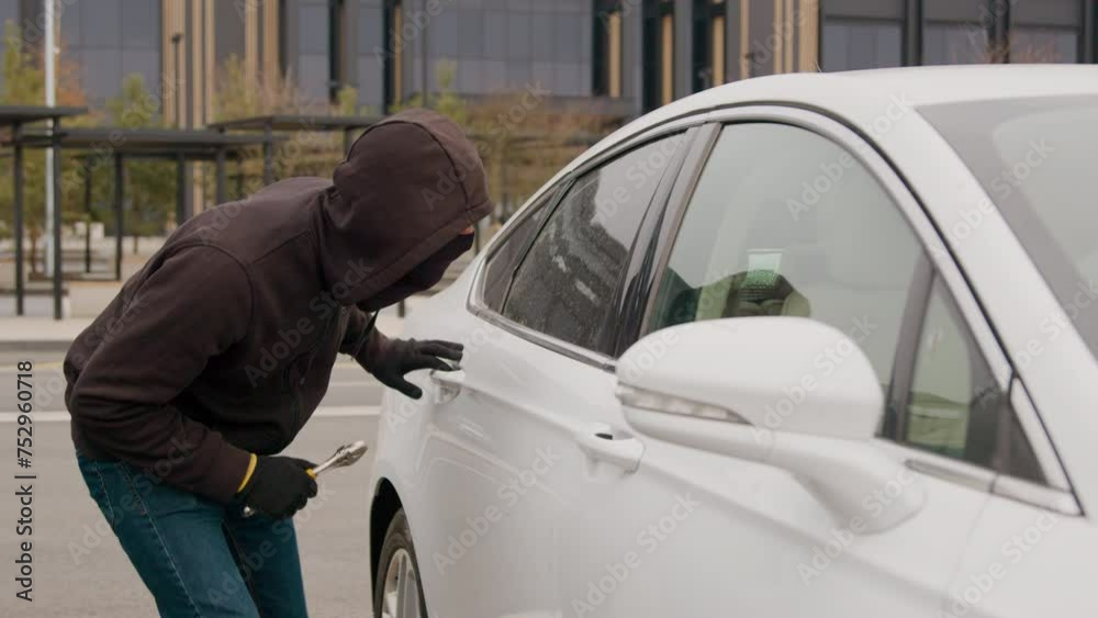 In a parking lot, a thief with a key in hand shatters the window of a white car, initiating a daring act of auto theft in broad daylight, amidst the hustle and bustle of the surroundings In a parking lot, a thief with a key in hand shatters the window of a white car, initiating a daring act of auto theft in broad daylight, amidst the hustle and bustle of the surroundings