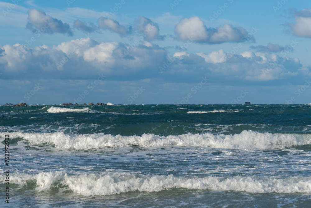 De petites vagues caressent le littoral breton sous un ciel bleu parsemé de quelques nuages, révélant la quiétude et la beauté tranquille de cette côte aux eaux turquoises.
