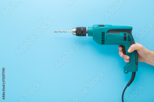 Young adult man hand holding and showing new dark black green professional electric hammer driver drill on light blue wall background. Pastel color. Closeup. Tool for repair work. Side view.