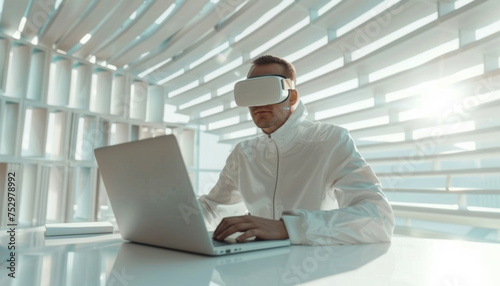 a man in virtual reality glasses works at a laptop in a white office.