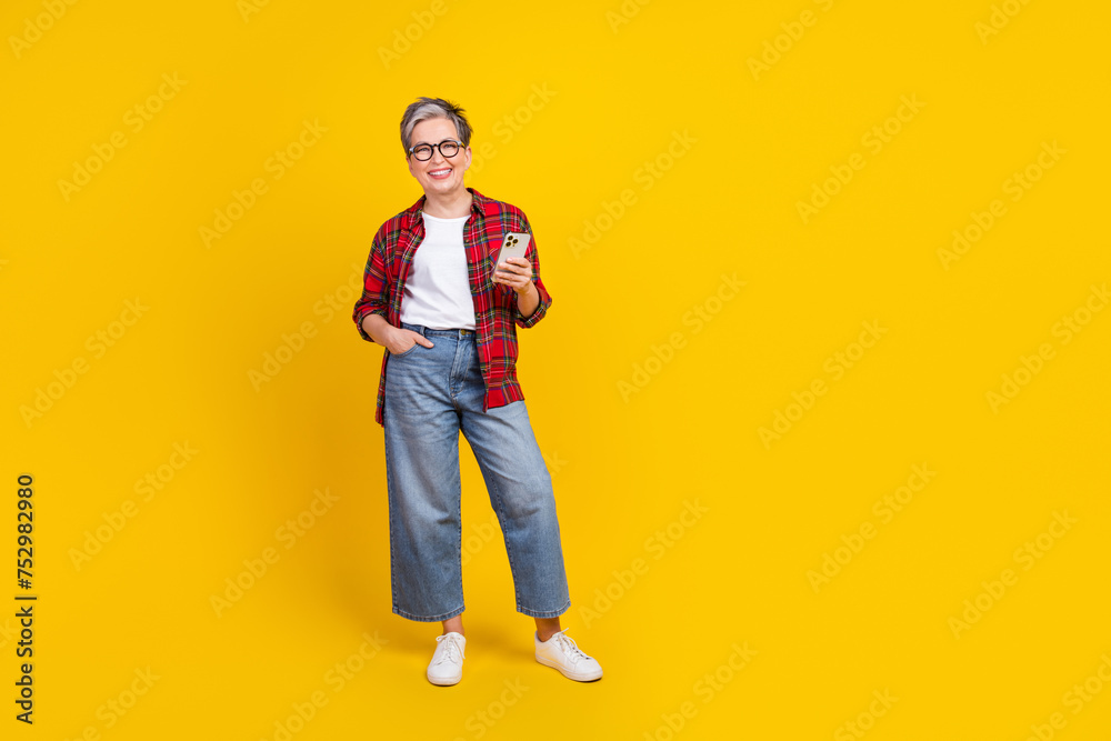 Full size photo of cheerful grandmother hold smart phone empty space ad isolated on yellow color background Full size photo of cheerful grandmother hold smart phone empty space ad isolated on yellow color background