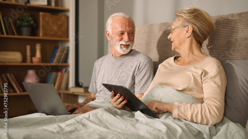 Smiling senior couple using a digital tablet and a laptop in their bed