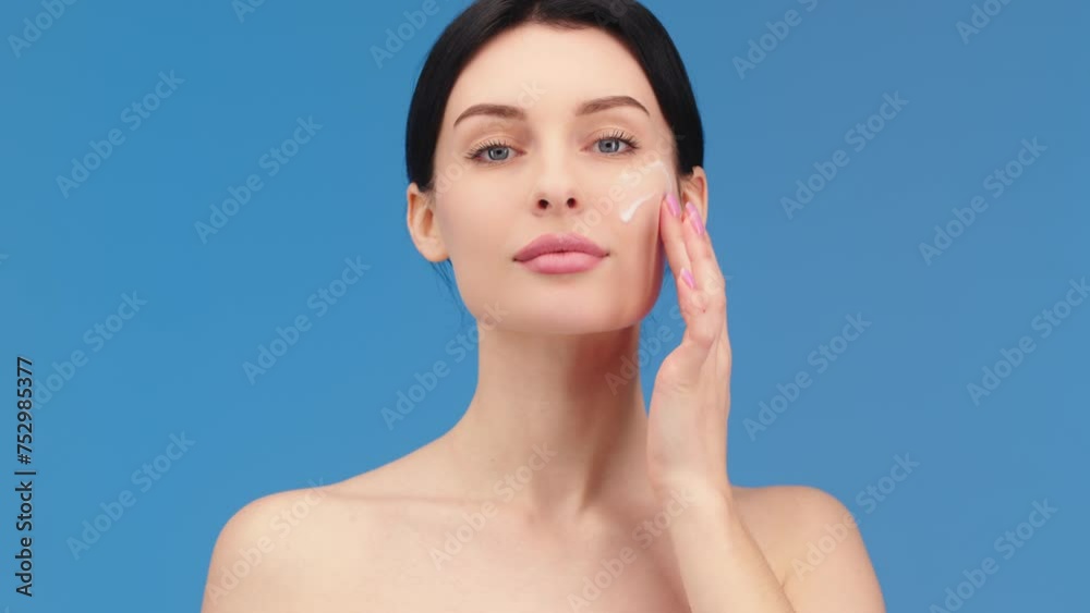 Face of a woman with blue eyes applying cream to her face and looking at the camera. Beautiful female model with perfectly clean fresh skin. Skin care or cosmetic advertising concept