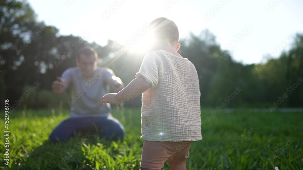 Fototapeta premium baby takes first steps in the park. happy family kid dream concept. dad teaches his son to walk takes the first steps lifestyle in the park on green grass. baby son taking first steps